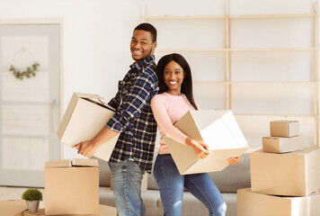 Cheerful black couple moving to new residence together, holding belongings on relocation day. Young African American family feeling happy about their rented place of living