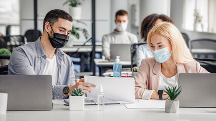 Obraz premium Social distance and work with documents in modern office. Millennial man and woman in protective masks in workplace with laptops and antiseptic, talk after quarantine covid-19, panorama, copy space