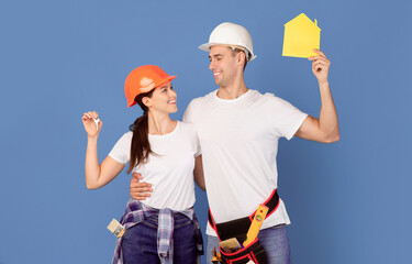Home Improvement. Happy Young Couple In Hardhats Holding Yellow Paper House Figure And Keys, Enjoying Making Renovation Together, Celebrating Buying Property, Standing Over Blue Background, Copy Space