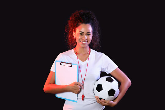 Female African-American soccer trainer with ball and clipboard on black background