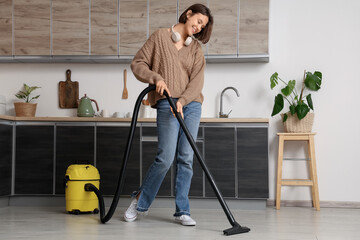 Beautiful young woman with modern vacuum cleaner and headphones cleaning floor in kitchen