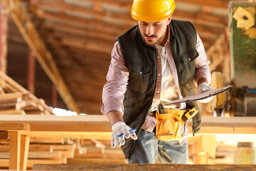 Young carpenter in hardhat with clipboard working at sawmill