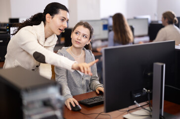 Young female teacher helping young female trainee work at computer in office