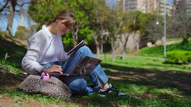 Young female student sitting on grassy park ground, studying intently using laptop and notebook, periodically writing notes while enjoying peaceful outdoor learning environment