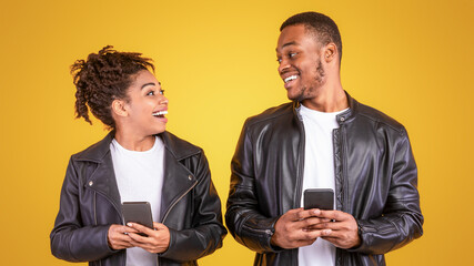 Cheerful African American Couple Using Smartphone Texting In Online Chat Standing Over Yellow Studio Background. Mobile Communication And Phone Applications Concept