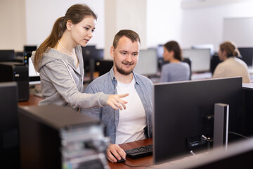 In classroom during computer courses, female teacher helps man student complete difficult task, looks at monitor screen and corrects work. Mentor comments on students work, gives hints during task.