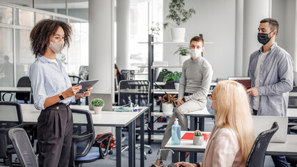 Corporate meeting and group work in modern company in office interior. African american woman manager in protective mask holding tablet, talking to workers keeping social distance during epidemic