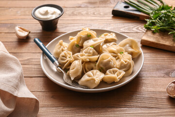 Plate of delicious boiled dumplings with parsley and bowl of sour cream on wooden background, closeup