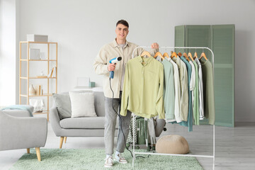 Young man with steamer and clothes on hanger at home