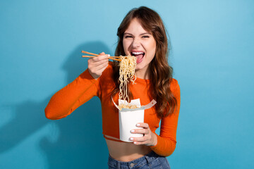 Young woman enjoying noodles with chopsticks against blue background in casual fashion, expressing happiness and fun