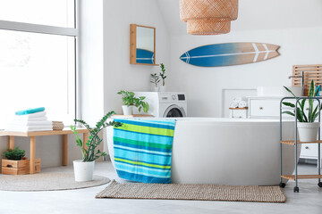 Interior of light bathroom with bathtub, washing machine, big window and surfboard