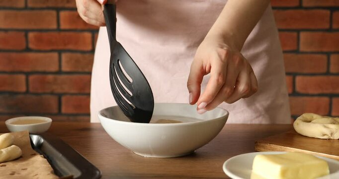 Woman putting uncooked pretzel into bowl of water at wooden table, closeup