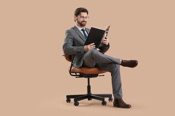 Handsome businessman reading magazine in chair on beige background