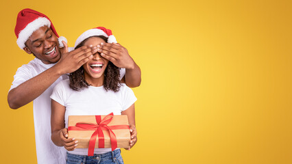 Surprise For Christmas. African Husband Congratulating Wife On Xmas With Wrapped Gift Box Standing On Yellow Studio Background, Covering Her Eyes. Winter Holidays And New Year Celebration. Panorama