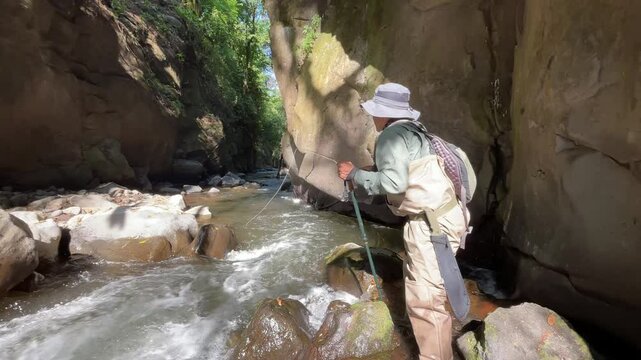 Wide shot of a fisherman throwing a line into a river, standing among rocks with morning light shining through the forest.