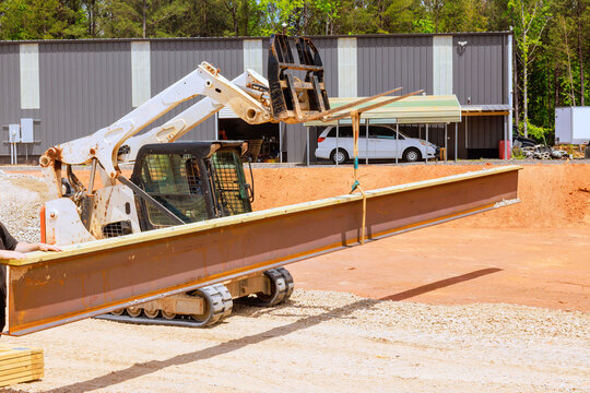 Construction worker operates forklift skid steer loader, moving large steel beam at building site