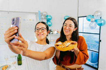 Two young Latin women, in a festively decorated office, take a selfie and hold a bowl of empanadas