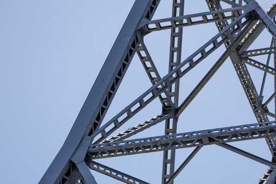 Network of steel beams forms a geometric pattern against a clear blue sky. Structure's angular lines and intricate metallic framework create a sense of precision in architectural design. - Powered by Adobe