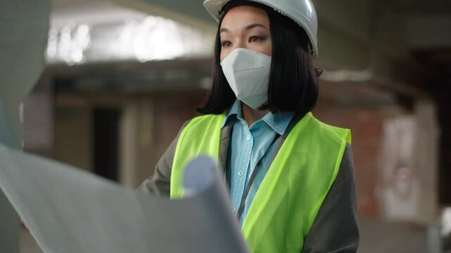 Asian Korean Chinese Japanese woman developer in safety mask hard hat female engineer builder architect inspector looking at building project plan construction girl checking blueprint industry worker