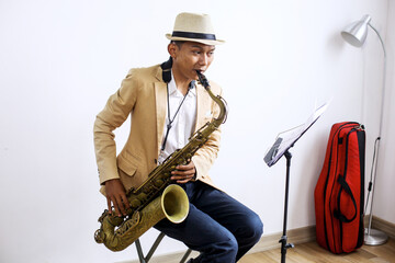 Naklejka premium Young Musician Man Blowing Saxophone While Sitting In Front Of Musical Stand At Home