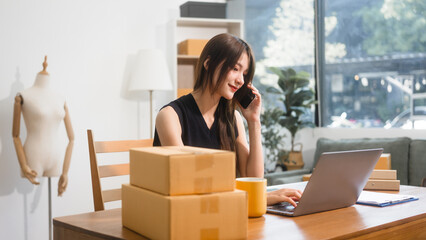 Happy Asian woman packing parcel boxes at a sofa table, smiling joyfully while checking online...