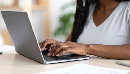 Fototapeta premium A focused woman with long brown hair sits at a wooden desk, typing intently on her sleek silver laptop, surrounded by colorful notebooks and a steaming coffee cup.