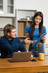 Caucasian man and an Asian woman sit at a table at home, engaged in online learning. They share...
