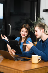 Caucasian man and an Asian woman sit at a table at home, engaged in online learning. They share...