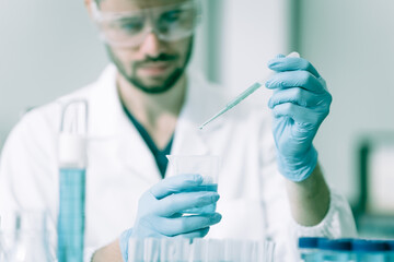 A white male scientist at a table, studying blue substances in test tubes and bottles, using a...