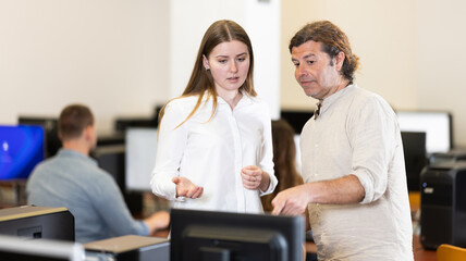 Man tutor and girl standing near computer, discussing educational process, progress and academic performance of students. Computer course worker and student are having nice conversation in classroom.