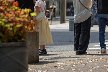 Casually dressed people stand at a street corner on a warm spring day, waiting to cross pedestrian crosswalk under bright daylight amidst urban surroundings.