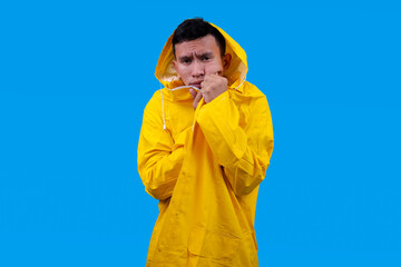 Young man wearing rain coat going cold due to low temperature isolated over blue background