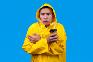 Young man in raincoat hood feeling cold while drink a hot coffee in cup isolated on blue background