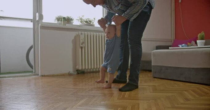 A father helps his baby take their first steps at home. The baby is learning to walk with the support of his loving parent.