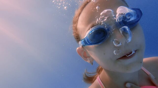 Close up portrait of a young girl expelling air from her nose, creating a stream of bubbles while swimming underwater in a pool, wearing blue swimming goggles