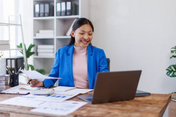 Young business asian woman typing on laptop holding papers preparing report analyzing work results,...