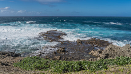 The waves of the turquoise ocean are raging and crashing against the coastal cliffs. White foam on the water. In the foreground are the volcanic rocks of the coast, green vegetation. Blue sky, clouds.