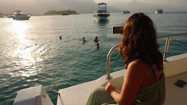 Female tourist capturing memorable moments of friends swimming in ocean waters during golden sunset, recording scenic seascape from boat stern using smartphone