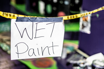 Handwritten wet paint warning sign hanging from yellow caution tape in neon painted room