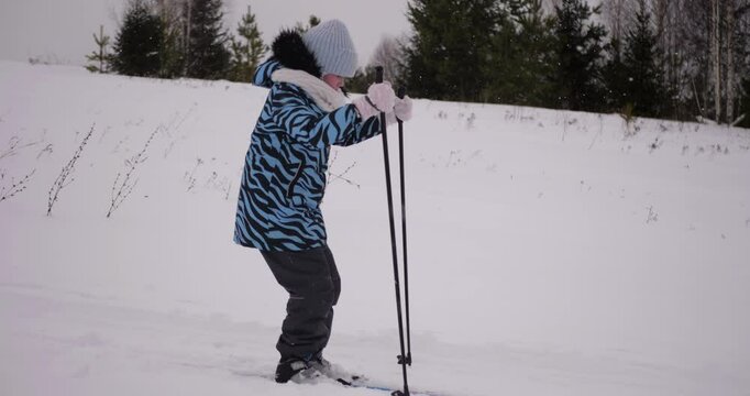 9-year-old girl carefully skiing down gentle slope on cross-country skis during snowfall, wearing winter clothes and ushanka hat