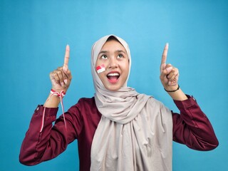 An enthusiastic Indonesian Muslim woman in hijab points upward with patriotic Red-and-White flag...