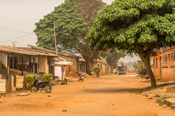Dusty rural street in Ouidah, Benin lined with modest houses, lush trees, motorbikes, and local market stalls, capturing authentic West African life.
