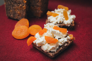 Slices of homemade healthy bread with fresh cheese and dried apricots, with whole dried apricots fruits on dark red background close-up