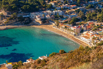 Fototapeta premium Clear turquoise water laps against the golden sand of El Portet beach in Moraira, Spain, as palm trees and Mediterranean houses line the peaceful bay in this elevated coastal view