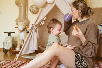 Mother and her baby boy playing at home. Mom is trying to eat her cookie and drink coffee in the morning.