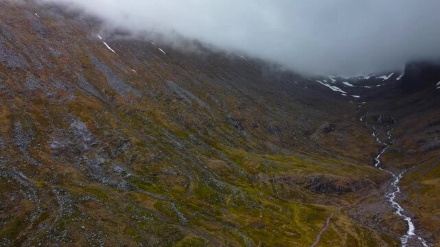 Gorgeous aerial drone footage flying above and over a small fast flowing white colored river or river rapis in scottish highlands mountains or hills. White fluffy clouds are low to the ground hovering