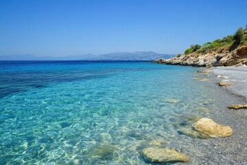 Fototapeta premium Crystal clear Aegean Sea waters gently lap against a rocky shore under a bright blue sky, Aegean sea coast cristal sea water background and clear