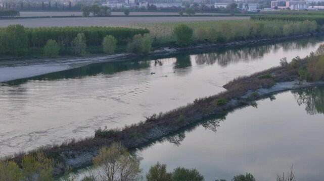 Distant rowers cut through calm Po River waters beside sandbars and tree-lined Maginot Budrio wetlands under warm sunset glow captured in real time