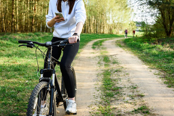 Young woman riding a bicycle and holding a phone in her other hand in the forest at sunset.