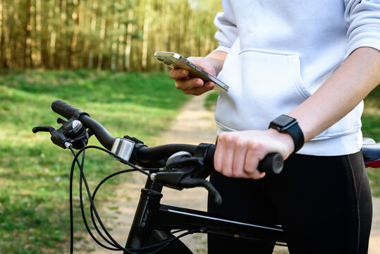 Close-up of a female cyclist's hand on the handlebars of a mountain bike and holding a phone in her other hand outdoors.
